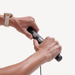 BodyFit — Black — Two hands gripping the black cylindrical device with silver buttons, cable attached, clean white background — close-up, two hands, cable