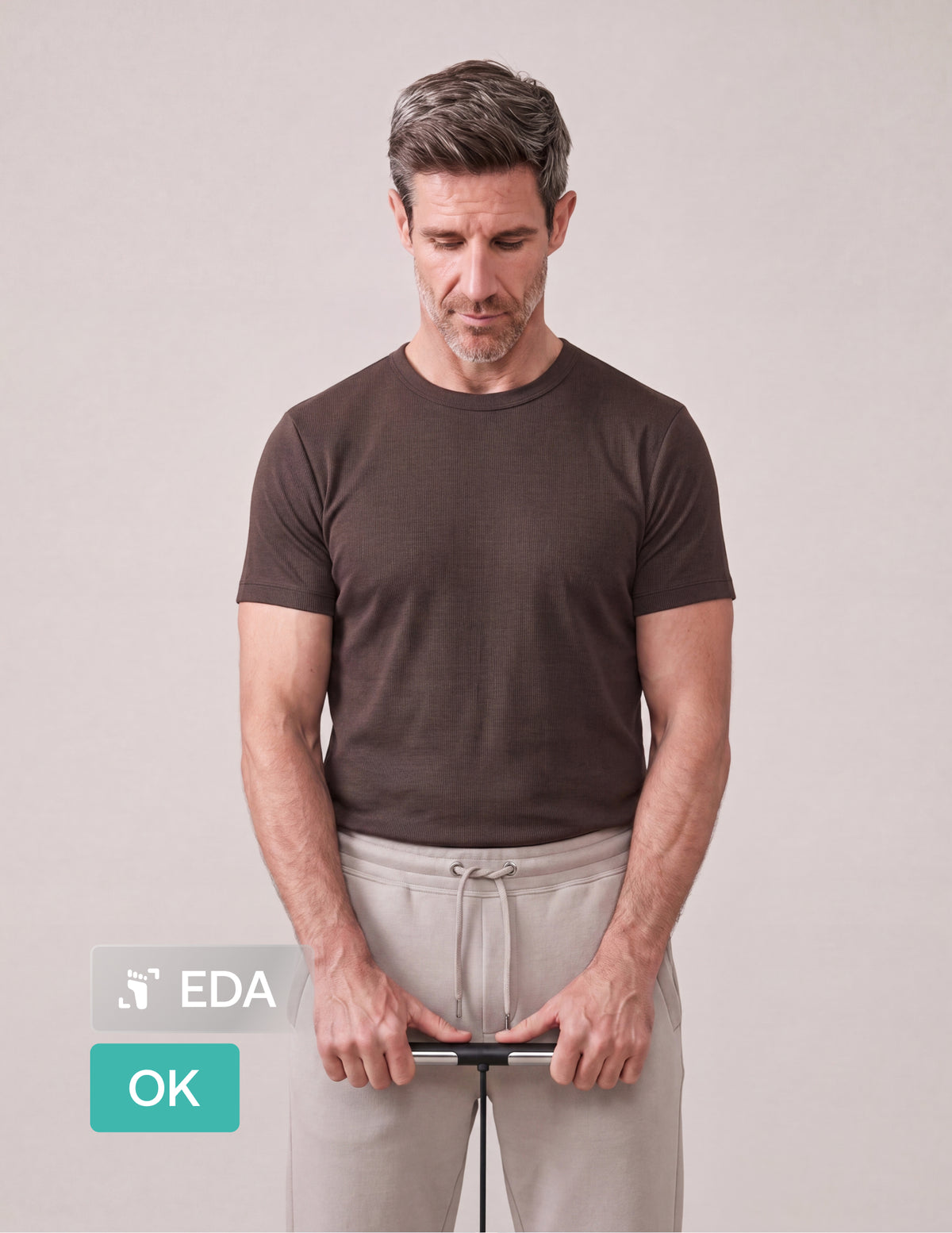 Withings Body Scan — Noir — Homme debout tenant la poignée de la balance, vue de face, t-shirt marron, fond neutre — poignée, corps entier, intérieur