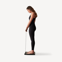Body Scan — Black — Woman standing side profile on the scale, holding measurement handle, black bodysuit — side view, full body, handheld sensors