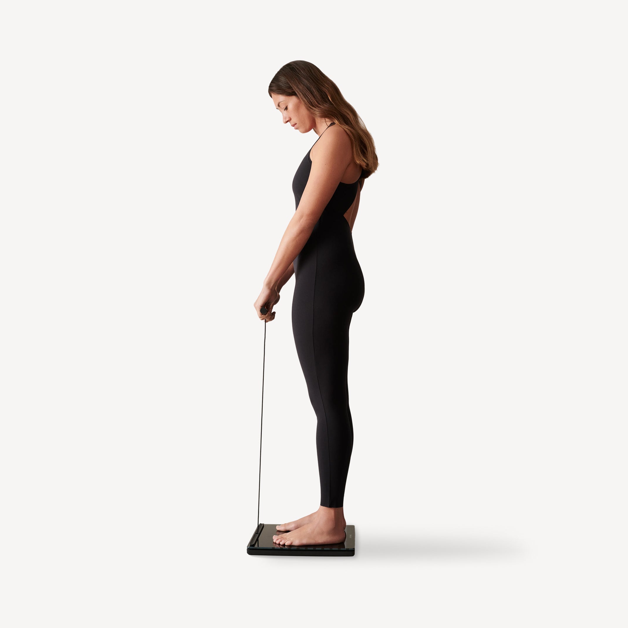 Body Scan — Black — Woman standing side profile on the scale, holding measurement handle, black bodysuit — side view, full body, handheld sensors