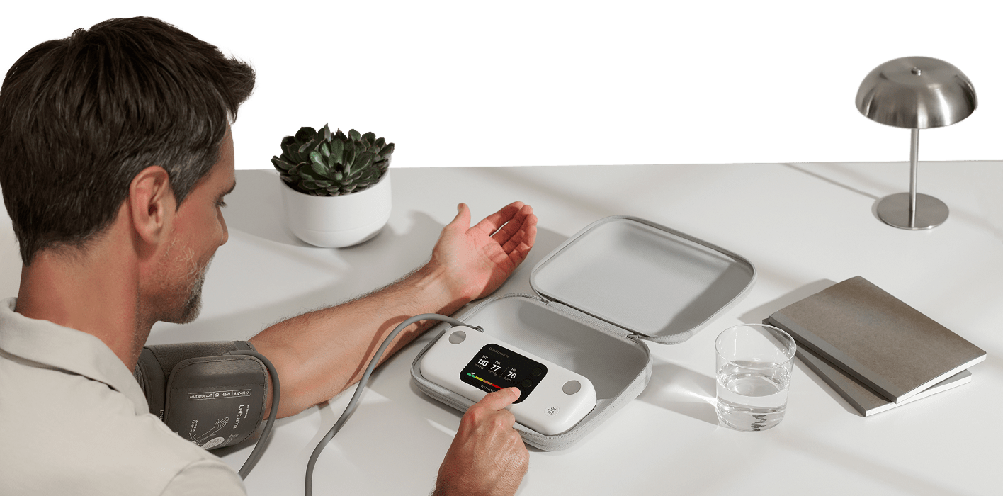 BPM Vision — bpm-vision — Man taking blood pressure reading, cuff on left upper arm, device on white desk displaying 116/77/76, indoor setting — desk, indoor, close-up, measurement