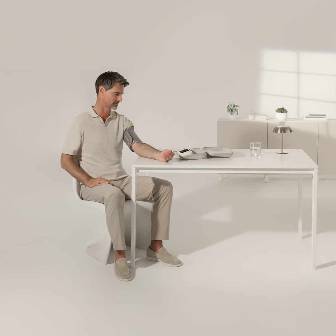 BPM Vision — Connected Blood Pressure Monitor — Man seated taking left-arm blood pressure measurement at white desk, minimal bright interior setting — blood pressure, left arm, seated, white desk