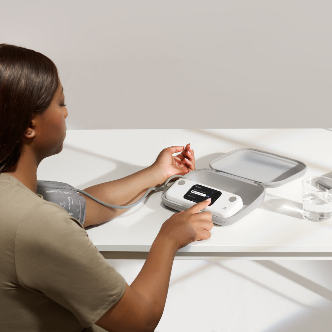 BPM Vision — Connected blood pressure monitor — Woman seated at a white table, cuff on left arm, pressing the device resting in its open case — table, natural light, indoor, three-quarter view
