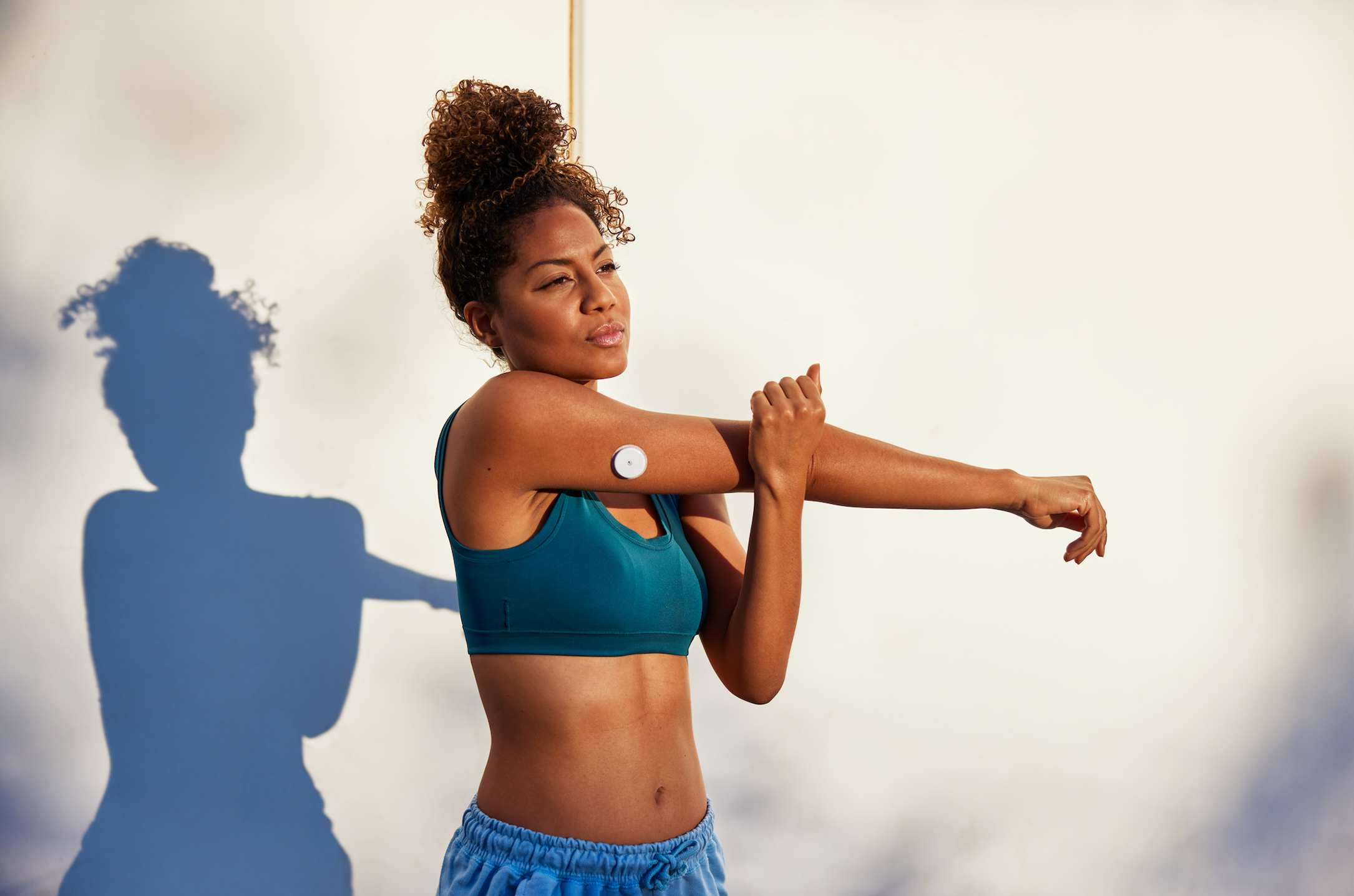 Lingo — Continuous glucose monitor — Woman in sportswear stretching arm with white sensor patch visible on upper arm, light background with blue shadow — sport, arm, sensor, stretch