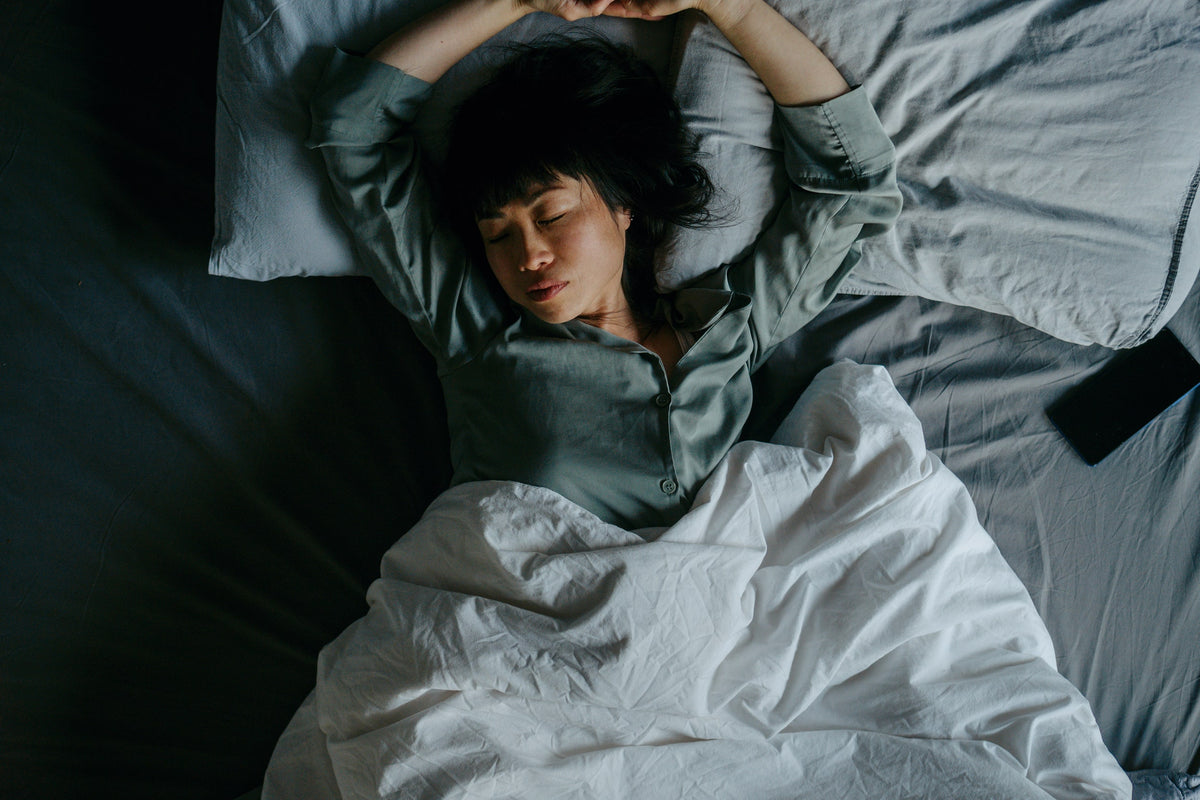 Withings — Press — Woman lying in bed seen from above, arms raised, grey and white sheets, dim lighting — sleep, relaxation, indoors
