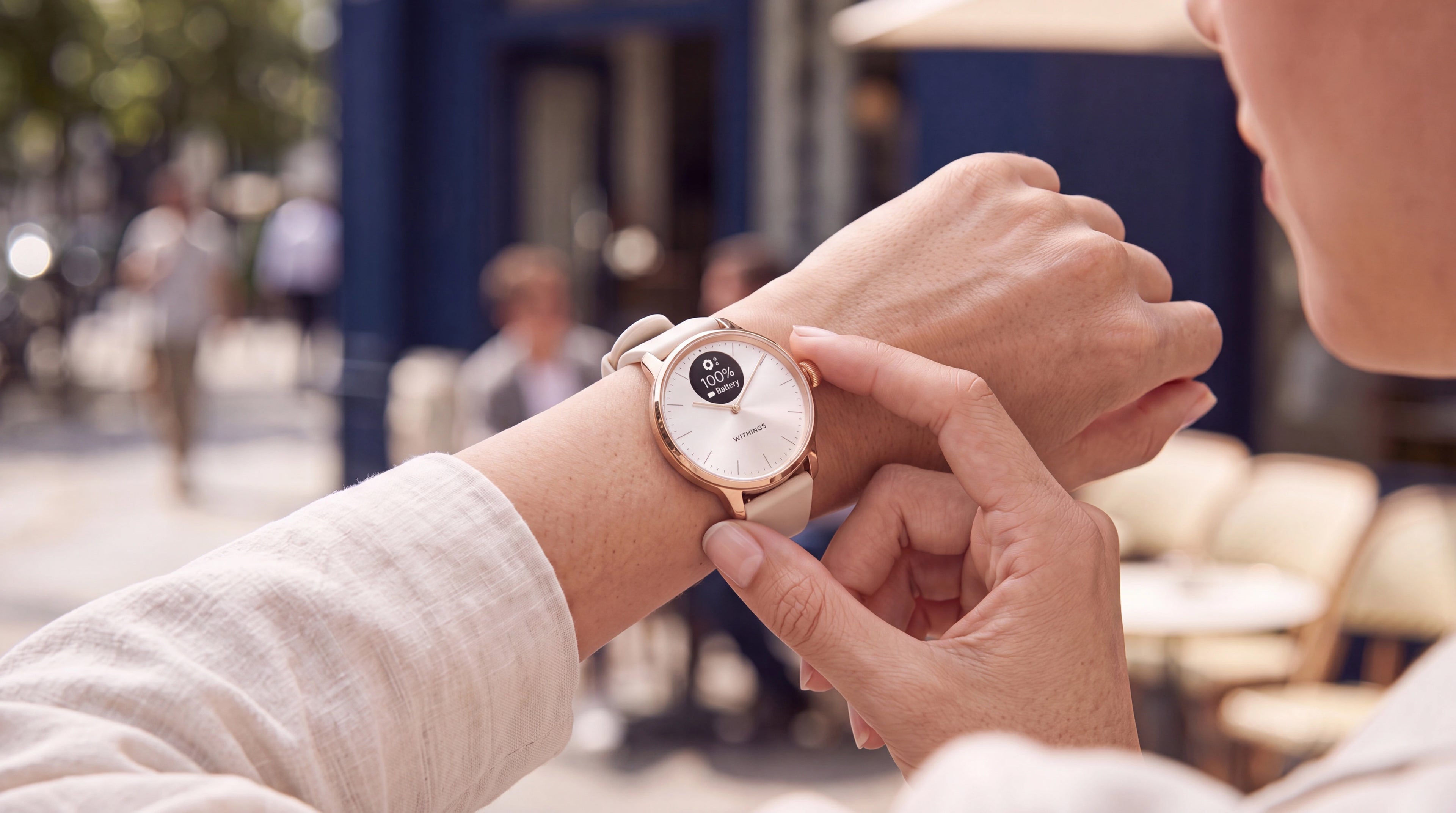 ScanWatch Light — Rose Gold 37mm White — Woman checking watch on wrist at urban outdoor terrace, battery display at 100%, beige strap — close-up, outdoor, wrist